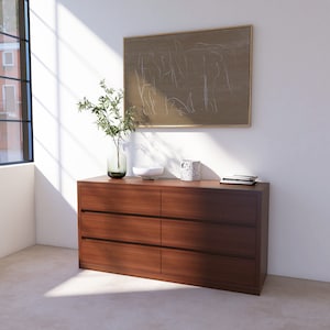 May include: A dark brown wooden dresser with six drawers. The dresser has a simple design with no visible hardware. The dresser is sitting on a light brown floor in front of a white wall. There is a framed picture hanging on the wall above the dresser.