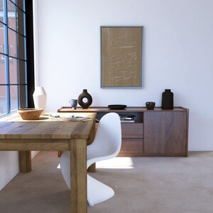 May include: A white modern chair sits at a wooden table in a minimalist room. A sideboard with two drawers and a cabinet door is behind the chair. A framed abstract art print hangs on the wall above the sideboard.