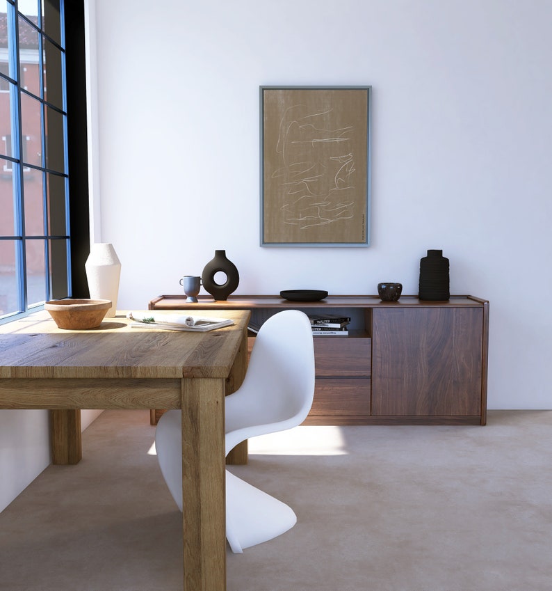 May include: A white modern chair sits at a wooden table in a minimalist room. A sideboard with two drawers and a cabinet door is behind the chair. A framed abstract art print hangs on the wall above the sideboard.