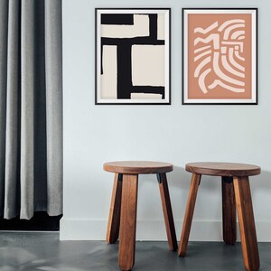 May include: Two wooden stools with a round seat and four legs. The stools are in front of a white wall with two framed abstract art prints. One print is black and white, and the other is peach and white.