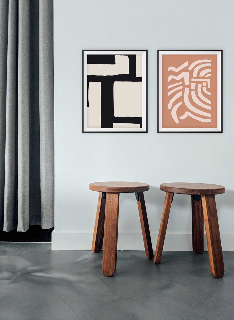 May include: Two wooden stools with a round seat and four legs. The stools are in front of a white wall with two framed abstract art prints. One print is black and white, and the other is peach and white.