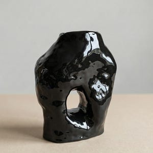 Puede incluir: Un jarrón de cerámica negro brillante con una forma orgánica y escultural. El jarrón tiene una gran abertura irregular y una superficie lisa y reflectante. El diseño es moderno y minimalista, adecuado para la decoración del hogar.