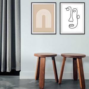May include: Two wooden stools with a round top and four legs. The stools are brown and have a natural wood finish. The stools are in front of a white wall with two framed prints. The print on the left is a beige background with a white abstract arch design. The print on the right is a white background with a black line drawing of a face.