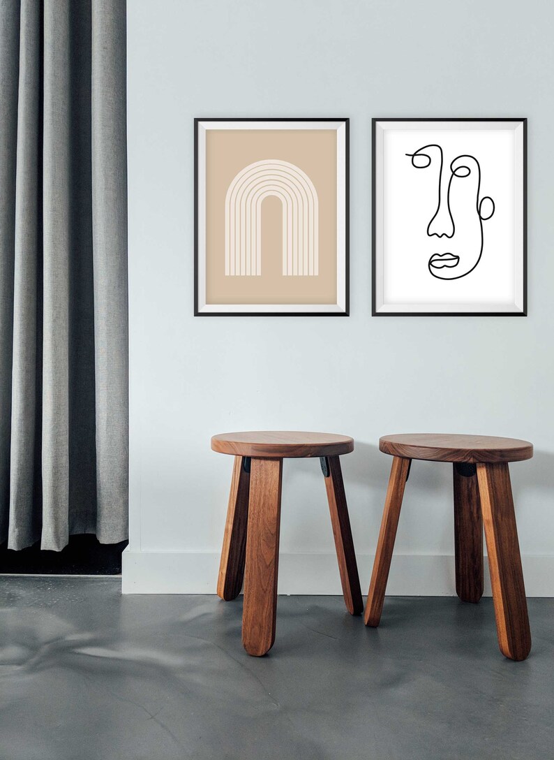 May include: Two wooden stools with a round top and four legs. The stools are brown and have a natural wood finish. The stools are in front of a white wall with two framed prints. The print on the left is a beige background with a white abstract arch design. The print on the right is a white background with a black line drawing of a face.