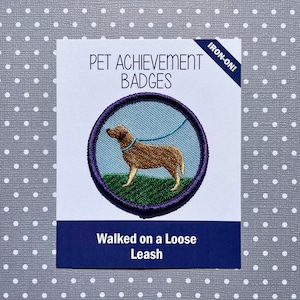 Puede incluir: Insignia de logro para mascotas para planchar con un perro marrón caminando con una correa azul. La insignia es morada con un fondo de hierba verde y el texto "Walked on a Loose Leash".