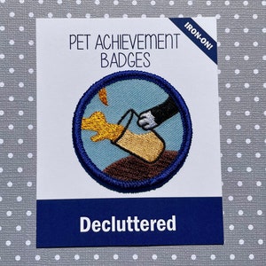 Puede incluir: Insignia de logro para mascotas para planchar con un fondo azul y una pata que llega a una taza amarilla. El texto "Decluttered" está impreso debajo de la insignia.