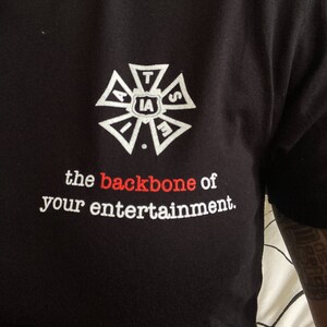 May include: Black t-shirt featuring a white and red emblem and text. The emblem has the letters "IA" at its centre. The text below reads "the backbone of your entertainment."