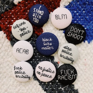 Könnte beinhalten: Eine Sammlung gestickter runder Anstecknadeln mit verschiedenen Phrasen und Slogans. Die Anstecknadeln sind weiß, marineblau und schwarz, mit Text in Schwarz und Weiß. Zu den Phrasen gehören "I STAND WITH YOU", "BLM", "DON'T SHOOT" und "ACAB".