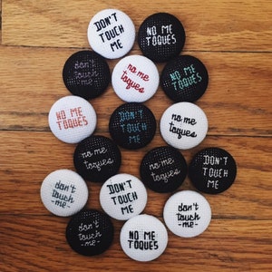 May include: A collection of round, embroidered pins with phrases like "Don't Touch Me" and "No Me Toques" in various colors. The pins are white and black, with text in black, white, red, and teal. The pins are arranged on a wooden surface.