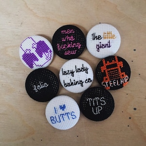 May include: A collection of handmade, embroidered round pins with various text and graphic designs. The pins feature phrases like "men who f*cking sew," "lazy lady baking co," and "I love butts." Colors include black, white, purple, orange, and blue.