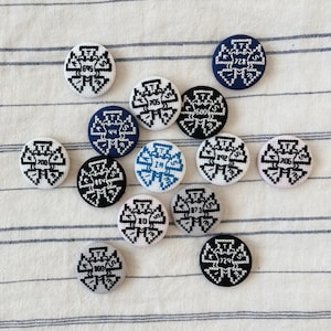 Puede incluir: Colección de botones redondos bordados en varios colores, incluyendo blanco, azul marino, negro y rosa. Cada botón presenta un diseño único con números y letras. Los botones están dispuestos sobre una tela a rayas.