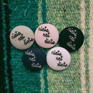 May include: Five round fabric buttons in various colors: light gray, pink, black, navy blue, and white. Each button has the embroidered text "nists not diets" in black cursive font. The buttons are arranged on a green and white striped surface.