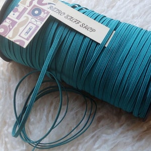 Op de afbeelding: Een spoel met turquoise elastisch koord met een label van "Retro Stuff Shop". Het koord is strak op een zwarte spoel gewikkeld, met wat koord afgewikkeld. De achtergrond is zacht en wit.
