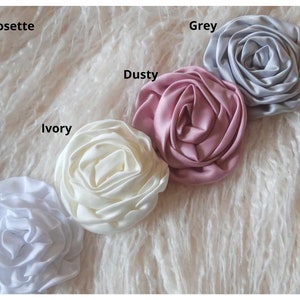 May include: Five 3-inch satin rosettes in different colors: white, ivory, dusty rose, grey, and white.  The rosettes are arranged in a row on a white fluffy surface.