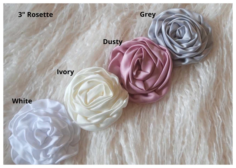 May include: Five 3-inch satin rosettes in different colors: white, ivory, dusty rose, grey, and white.  The rosettes are arranged in a row on a white fluffy surface.