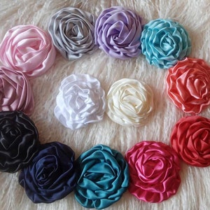 May include: A collection of 12 handmade fabric flower embellishments in various colors, including pink, purple, blue, white, cream, black, navy, teal, and red. The flowers are arranged in a circle on a white fluffy surface.