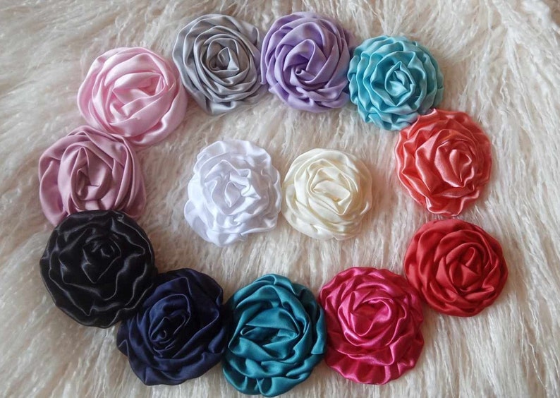 May include: A collection of 12 handmade fabric flower embellishments in various colors, including pink, purple, blue, white, cream, black, navy, teal, and red. The flowers are arranged in a circle on a white fluffy surface.