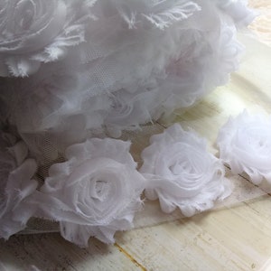 Peut inclure: Des embellissements floraux en tissu blanc avec une texture plissée. Les fleurs sont disposées sur un fond en maille blanche.