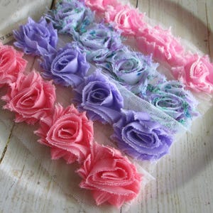 May include: A trim featuring rows of fabric roses in shades of pink, lavender, and blue attached to a white mesh backing. The roses have frayed edges and a layered construction. A small white tag with the text Retro Stuff Shop is visible.