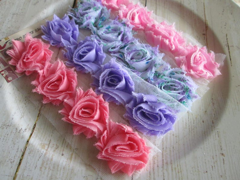 May include: A trim featuring rows of fabric roses in shades of pink, lavender, and blue attached to a white mesh backing. The roses have frayed edges and a layered construction. A small white tag with the text Retro Stuff Shop is visible.
