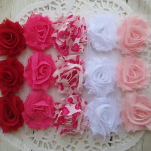 May include: A collection of fifteen fabric flowers arranged in a grid of five columns and three rows. The flowers feature frayed edges and include solid red, solid hot pink, pink and white heart-patterned, solid white, and solid light pink.
