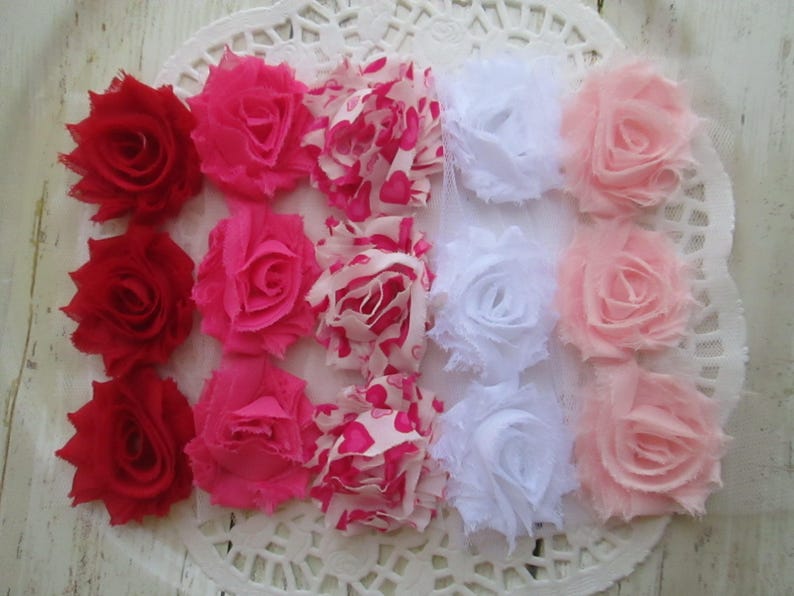 May include: A collection of fifteen fabric flowers arranged in a grid of five columns and three rows. The flowers feature frayed edges and include solid red, solid hot pink, pink and white heart-patterned, solid white, and solid light pink.