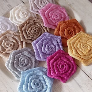 May include: Assortment of 12 fabric rose flower embellishments in various colors, including white, beige, light blue, lavender, pink, and yellow. The flowers are arranged in a circle on a white wooden surface.