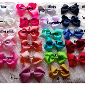 May include: A collection of colorful hair bows in various shades, including white, pink, blue, and red. Each bow is labeled with its color name, such as "White," "Navy," and "Red." The bows are arranged on a soft, white surface.