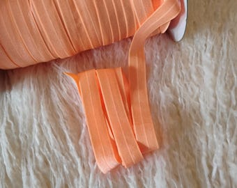 May include: A spool of orange elastic ribbon, partially unrolled, rests on a textured, off-white surface. The ribbon is a solid, warm orange color and appears to be pleated or folded. The image suggests a craft or sewing supply.
