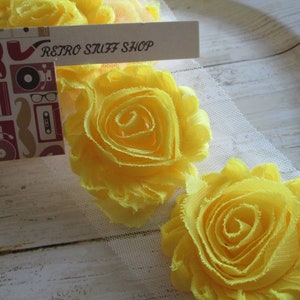 May include: Close-up of bright yellow fabric flowers with ruffled edges. A small tag with the text "RETRO STUFF SHOP" and retro-themed illustrations is placed near the flowers. The flowers are on a white mesh fabric.