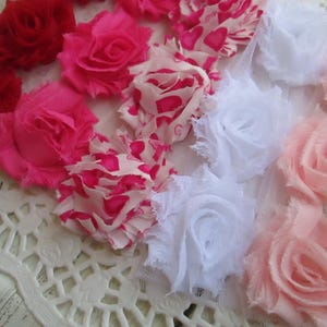 May include: A collection of fabric flowers in various shades of red, hot pink, light pink, and white, including some with heart patterns. These decorative rosettes feature frayed edges and layered petals, arranged in a cluster on a white lace base.