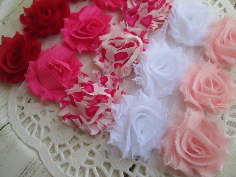 May include: A collection of fabric flowers in various shades of red, hot pink, light pink, and white, including some with heart patterns. These decorative rosettes feature frayed edges and layered petals, arranged in a cluster on a white lace base.