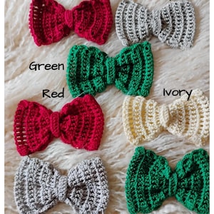 May include: A collection of handmade crochet bow hair accessories in various colors, including red, grey, green, and ivory. Each bow is labeled with its color name. The bows are displayed on a soft, textured surface.