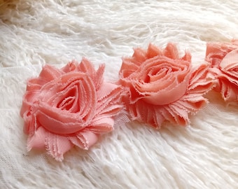 May include: Close-up of three peach-colored fabric flowers with ruffled edges. The flowers are arranged in a row on a white, textured background, creating a soft and delicate aesthetic. Ideal for crafting or embellishments.