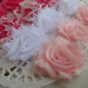 May include: A collection of fabric flowers featuring layered petals in shades of bright pink, white, and soft pale pink. The flowers are constructed from frayed-edge fabric and tulle, arranged in a cluster on a white lace-like base.