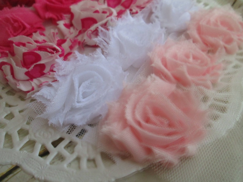 May include: A collection of fabric flowers featuring layered petals in shades of bright pink, white, and soft pale pink. The flowers are constructed from frayed-edge fabric and tulle, arranged in a cluster on a white lace-like base.