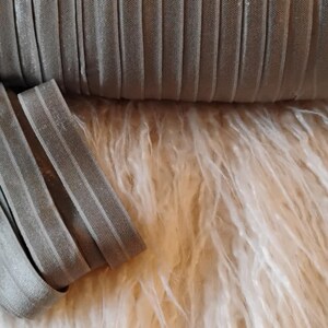 May include: A roll of gray, ribbed elastic ribbon with a folded section in the foreground. The ribbon is a muted gray color and appears to be made of a soft, flexible material. The background is a fluffy, white surface.