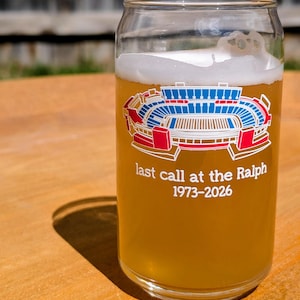 Può includere: Un bicchiere trasparente a forma di lattina di birra pieno di birra e schiuma. Il bicchiere presenta una grafica dello stadio in rosso, bianco e blu, con la scritta "last call at the Ralph 1973-2026". Il bicchiere è appoggiato su una superficie di legno.