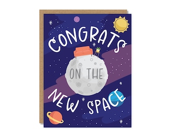 May include: A greeting card with a space-themed design. The card features a gray moon with a small orange couch and a potted plant. The words "CONGRATS ON THE NEW SPACE" are written in white script against a dark blue background with stars, planets, and a yellow sun.