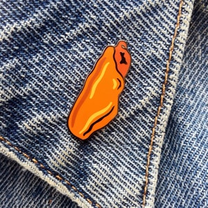 Op de afbeelding: Oranje en gele emaille pin in de vorm van een hotdog. De pin is op een blauw denim jack.