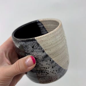 May include: Handmade ceramic tumbler with a unique design. The cup features a diagonal color block pattern in black, speckled gray, and off-white. The interior is black. A stylish and functional piece for beverages.