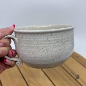 May include: A speckled, off-white ceramic mug with a rounded body and a curved handle. The mug has a wide opening and a simple, elegant design. The handle features a decorative swirl at the base. The mug is suitable for coffee, tea, or soup.