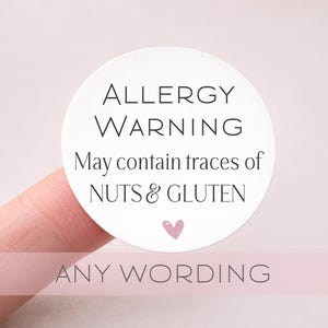 May include: A white, circular sticker with the words "ALLERGY WARNING" in a black, elegant font. Below, it reads "May contain traces of NUTS & GLUTEN" with a small pink heart. The bottom of the sticker reads "ANY WORDING".
