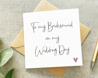 May include: A square white greeting card with black cursive text that reads To my Bridesmaid on my Wedding Day and a small pink heart icon in the bottom right corner. The card is placed on top of a tan envelope next to a gold pen.