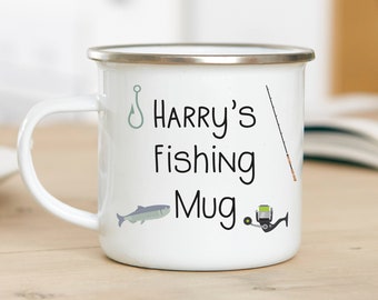May include: White enamel mug with a silver rim and a handle. The mug features the text "Harry's Fishing Mug" with illustrations of a fish hook, fish, fishing rod, and reel. The mug is on a wooden surface.