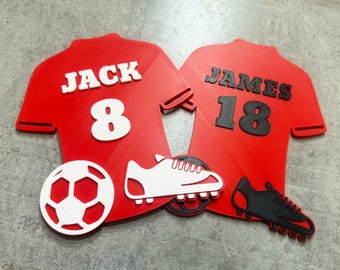 May include: Two red football shirts with white numbers and names. The shirts are cut out and have a white football and black football boots attached to them. The names on the shirts are "Jack 8" and "James 18".
