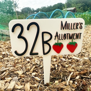 May include: A cream-coloured garden sign displaying the text "32B Miller's Allotment" in black and red. Two red strawberry illustrations with green leaves are featured. The sign is positioned in a garden plot.