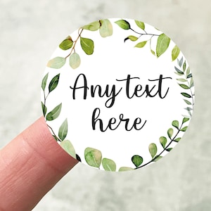 May include: A circular white sticker featuring the words "Any text here" in black script. The sticker is decorated with a watercolour design of green leaves and branches. A finger is holding the sticker.