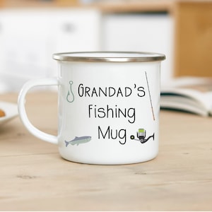May include: White enamel mug with a silver rim, displaying the text "Grandad's Fishing Mug". The design includes fishing-related illustrations: a fish, hook, rod, and reel. The mug sits on a light wooden surface, perfect for a fishing enthusiast.