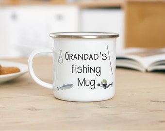 May include: White enamel mug with a silver rim, featuring the text "Grandad's Fishing Mug" in black script. The mug is decorated with fishing-themed illustrations, including a fish, hook, fishing rod, and reel. The mug is on a light wood surface.